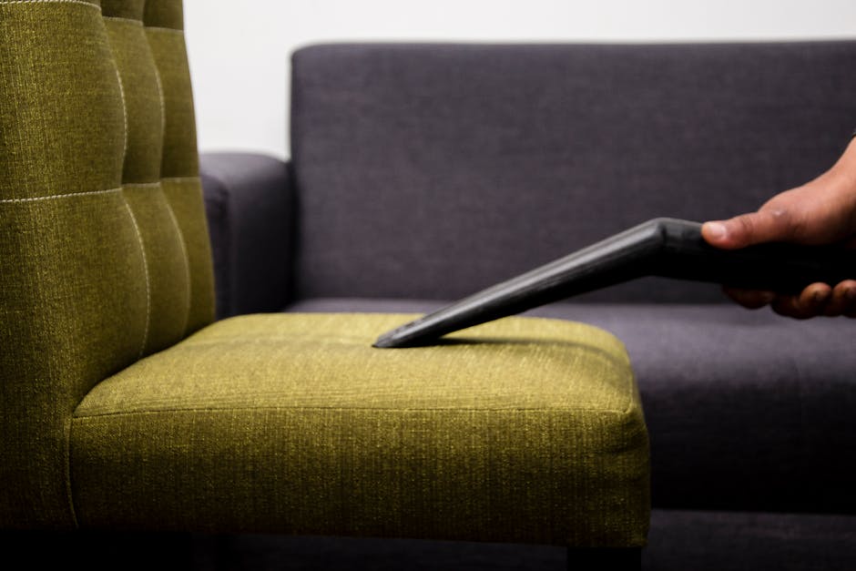A person using a handheld steam cleaning machine to deep clean a beige fabric upholstered armchair on a wooden floor. The machine's nozzle is pressed onto the fabric, releasing steam to remove dirt an